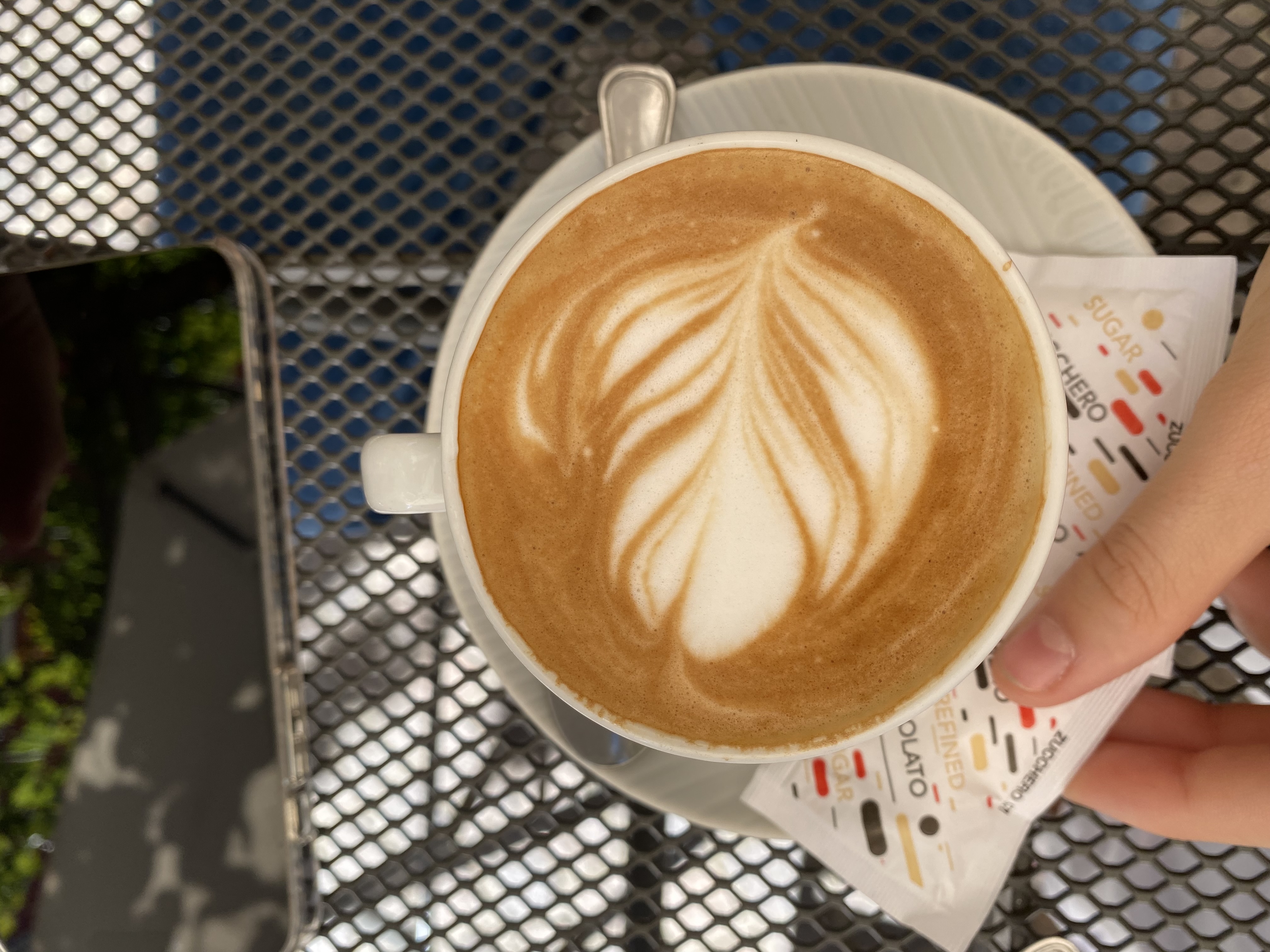Cappuccino with perfect leaf latte art, served on a Genova café terrace, metal mesh table in sunlight